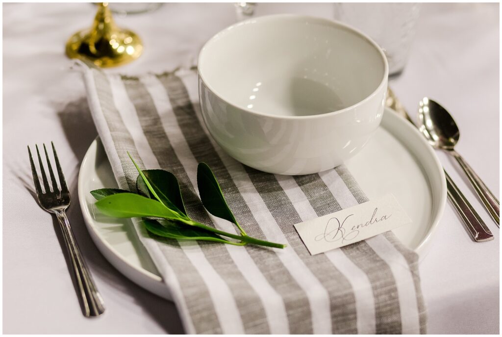 Wedding reception place setting with gray and white striped napkin, white plate and bowl, and sprig of greenery