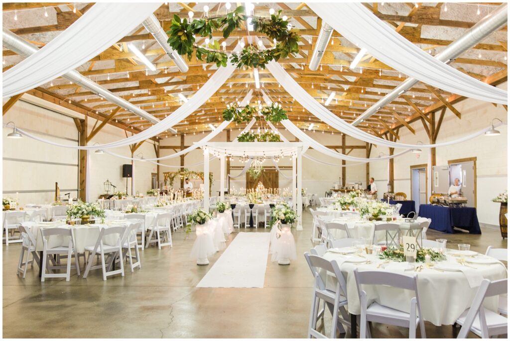 Beautiful wedding reception with hanging greenery, lights and white cloth. Tablescapes use white tablecloths, white florals and greenery.